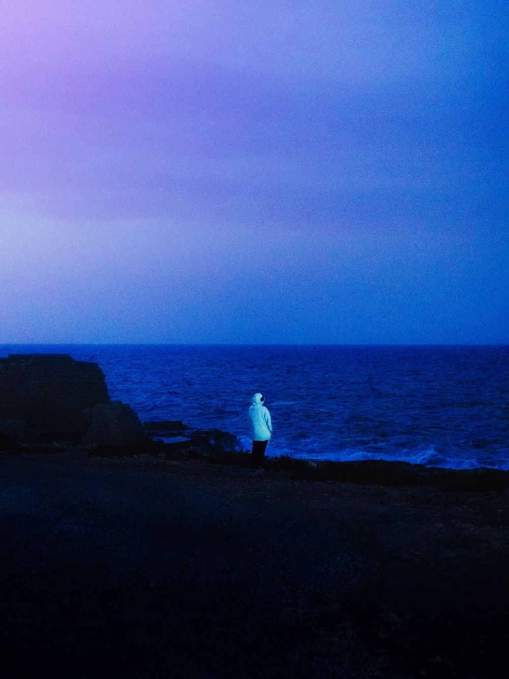 Silhouetted figure stands on rocky coastline against purple and blue twilight sky.