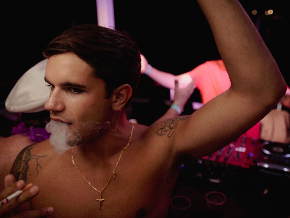 Shirtless performer with necklaces exhaling smoke under pink lighting at a club.