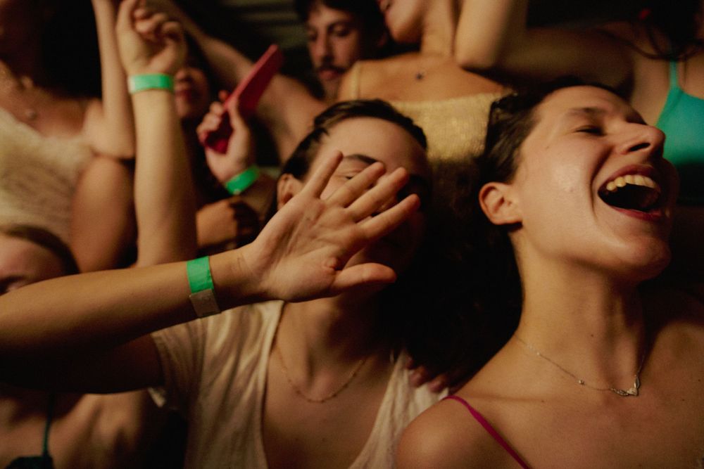 Crowd of concert-goers dancing enthusiastically with raised hands in a dark venue with neon lighting.