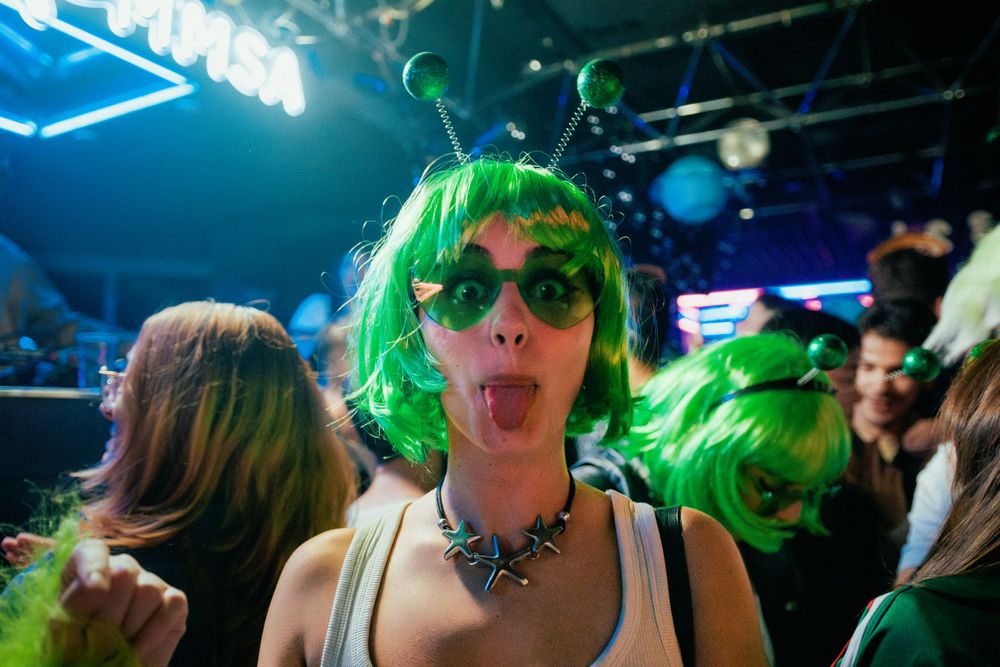 A party-goer with green hair and sunglasses sticks their tongue out at a neon-lit nightclub event.