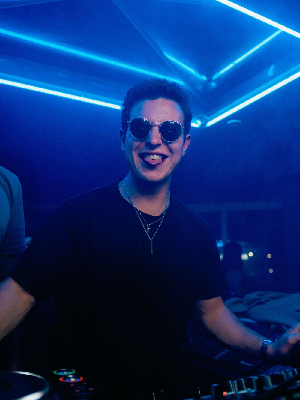 A DJ performs at a nightclub under vibrant blue neon lights wearing sunglasses and a black shirt.