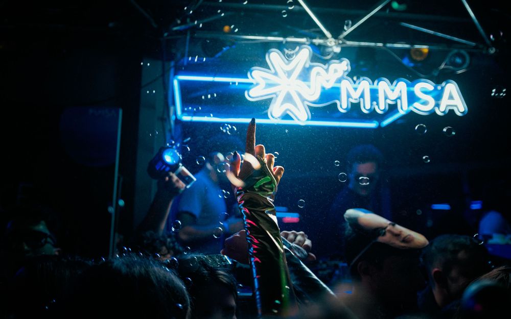 A performer on stage illuminated by blue lighting under a neon MMSA sign in a nightclub atmosphere.