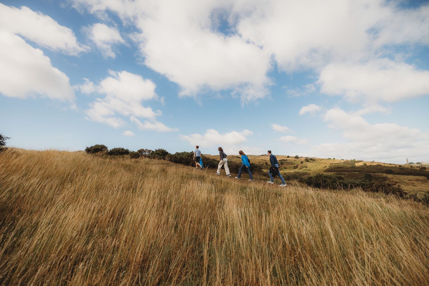 A panoramic view of golden grassy hills with people hiking along the ridgeline under a partly cloudy sky.