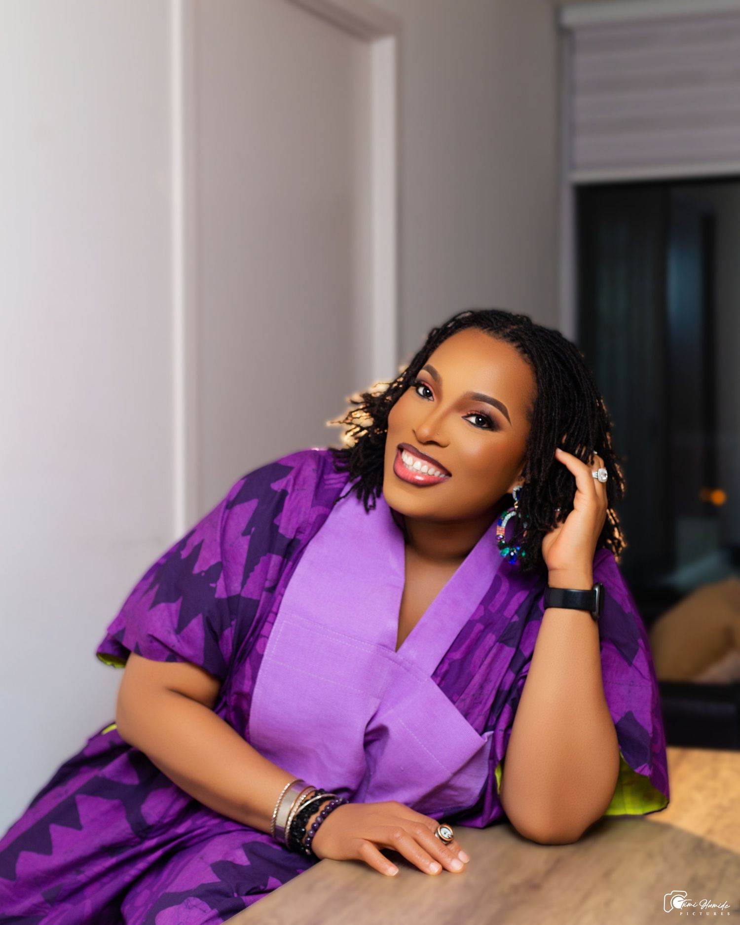 A woman wearing a vibrant purple outfit with gold accessories lounges on a couch in an elegantly lit room.
