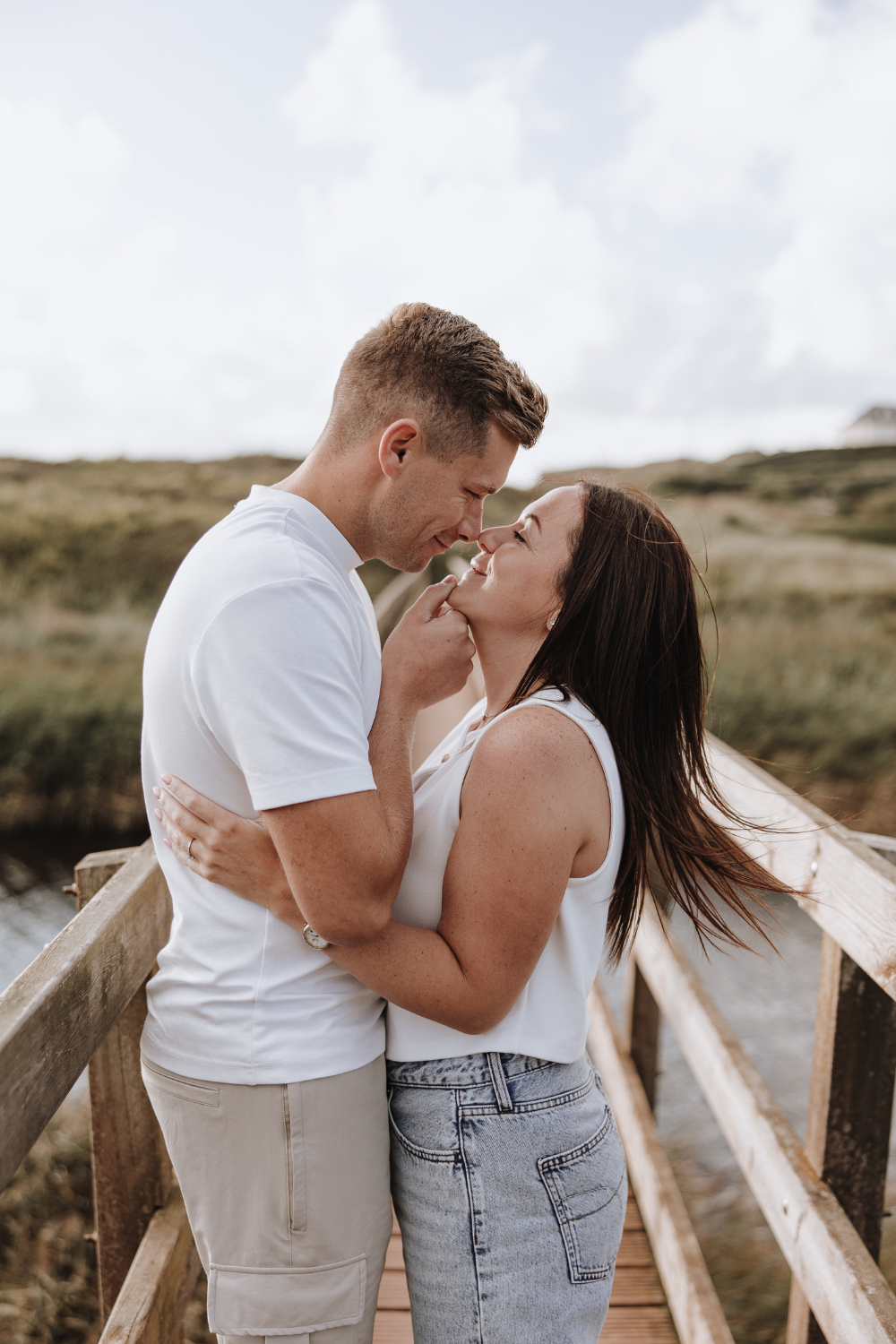 Romantic couple embracing and almost kissing on a wooden bridge outdoors, wearing casual white tops and jeans.