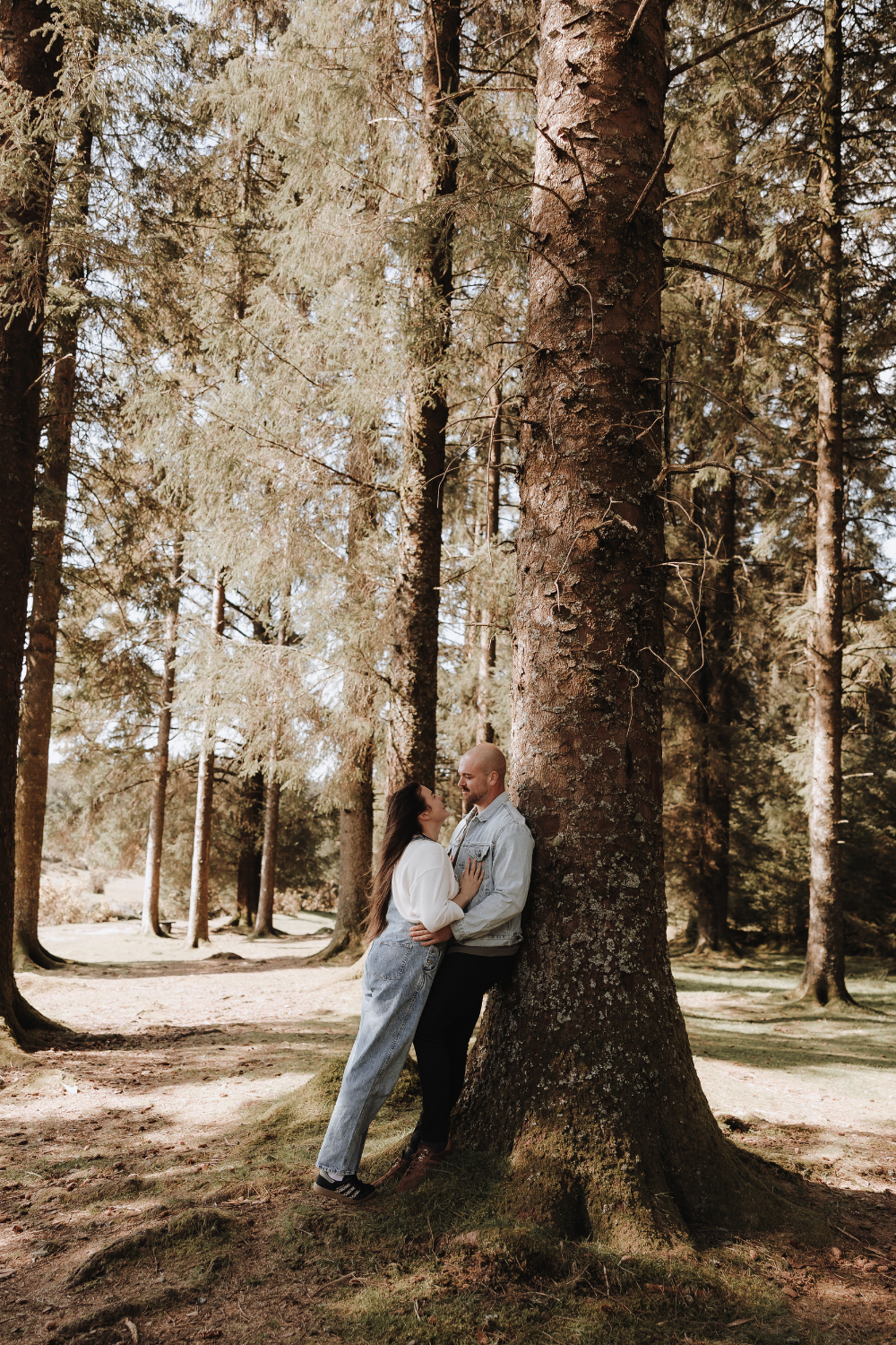 Couple leaning against a large tree trunk in a sunlit pine forest, sharing a romantic moment outdoors.