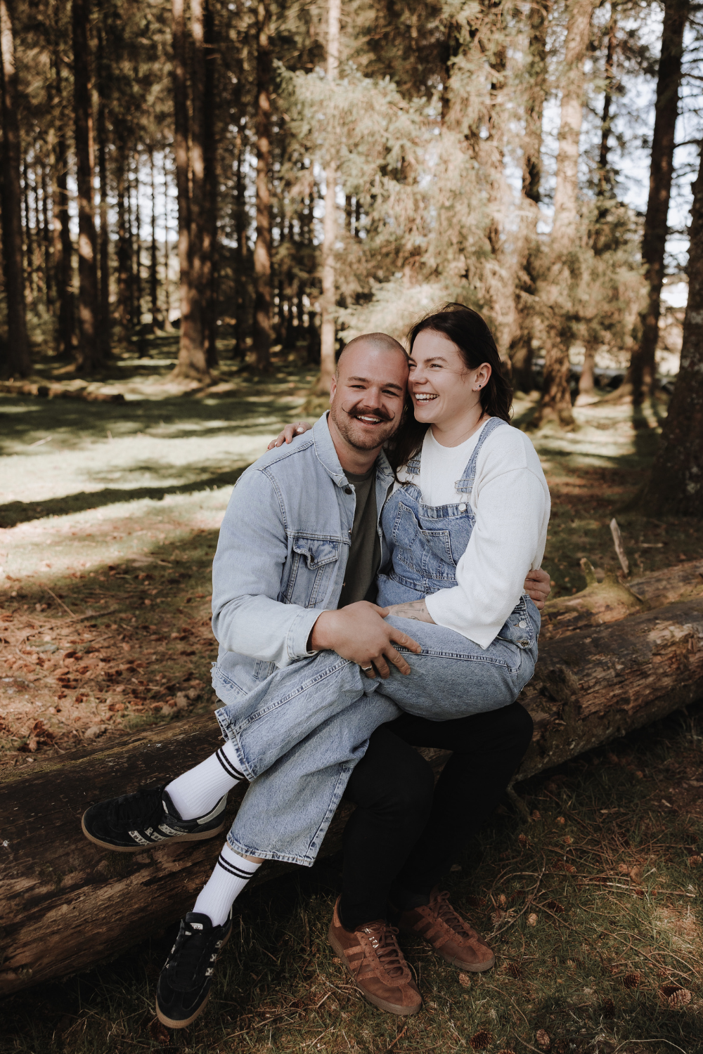 Happy couple sitting on a log in a forest, smiling and embracing in casual denim outfits.