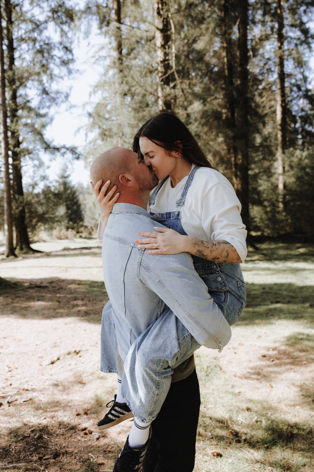 A bald man lifts and kisses a woman in denim overalls outdoors in a sunlit forest setting.