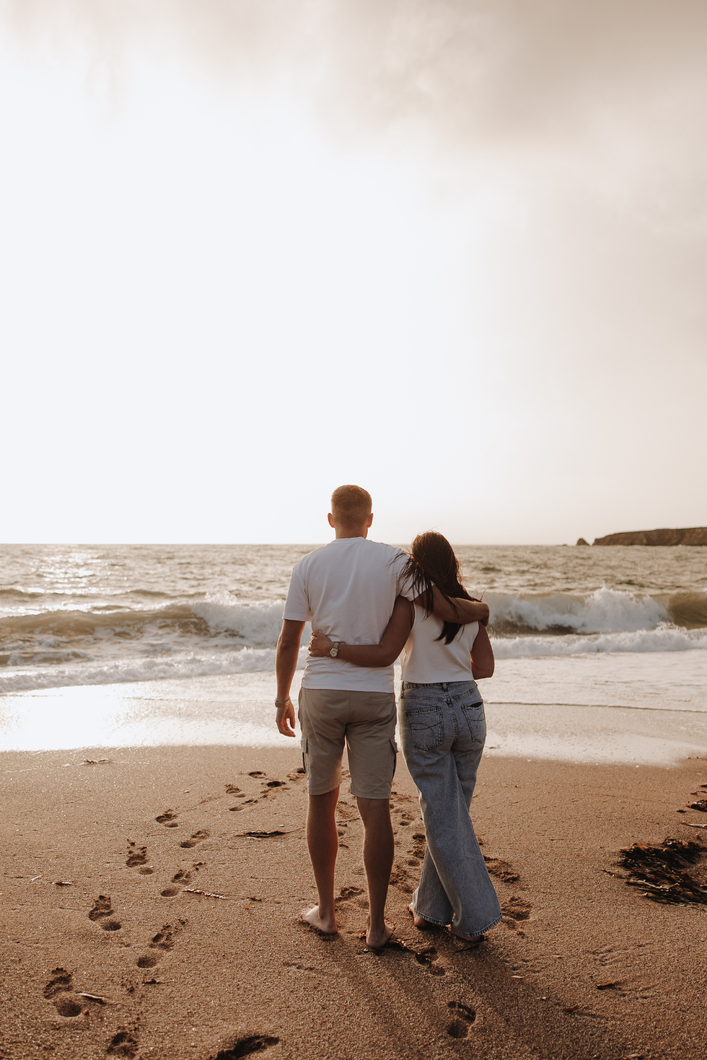 Couple embracing on sandy beach at sunset, walking barefoot with footprints in sand, ocean waves in background.