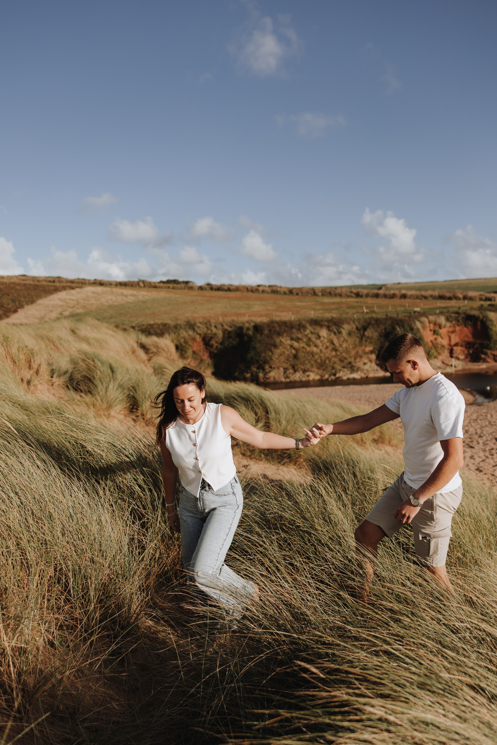 A couple holds hands while walking through tall grass dunes on a sunny coastal landscape.