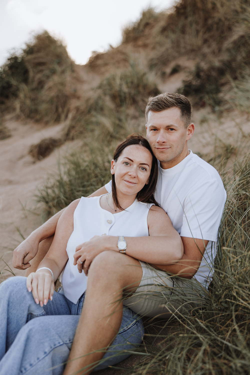 Couple sitting together on sandy beach dunes, woman in white top and jeans leaning against man in white t-shirt.