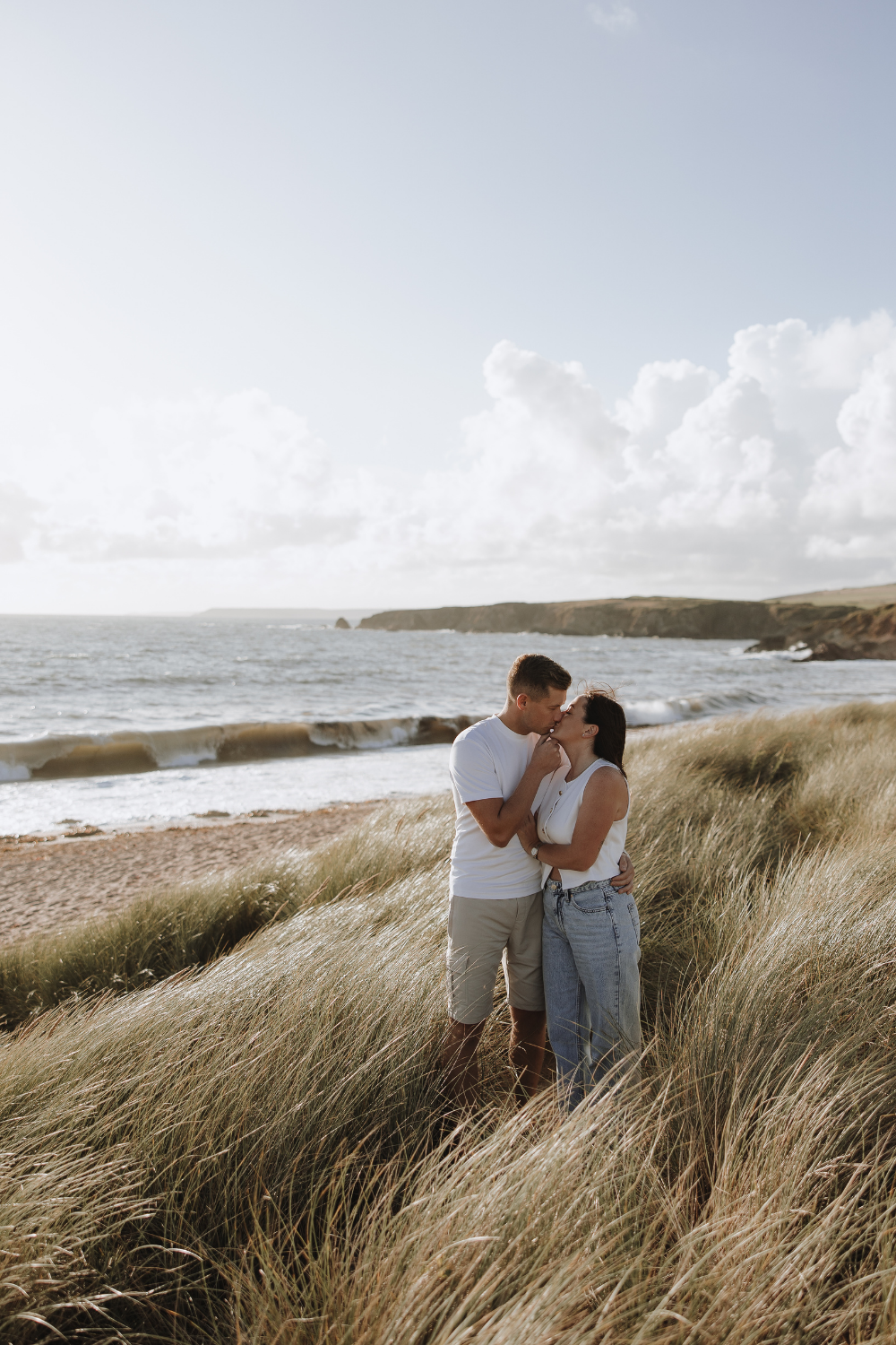 Couple kissing in coastal grass dunes with ocean waves and rocky shoreline in background on sunny day.