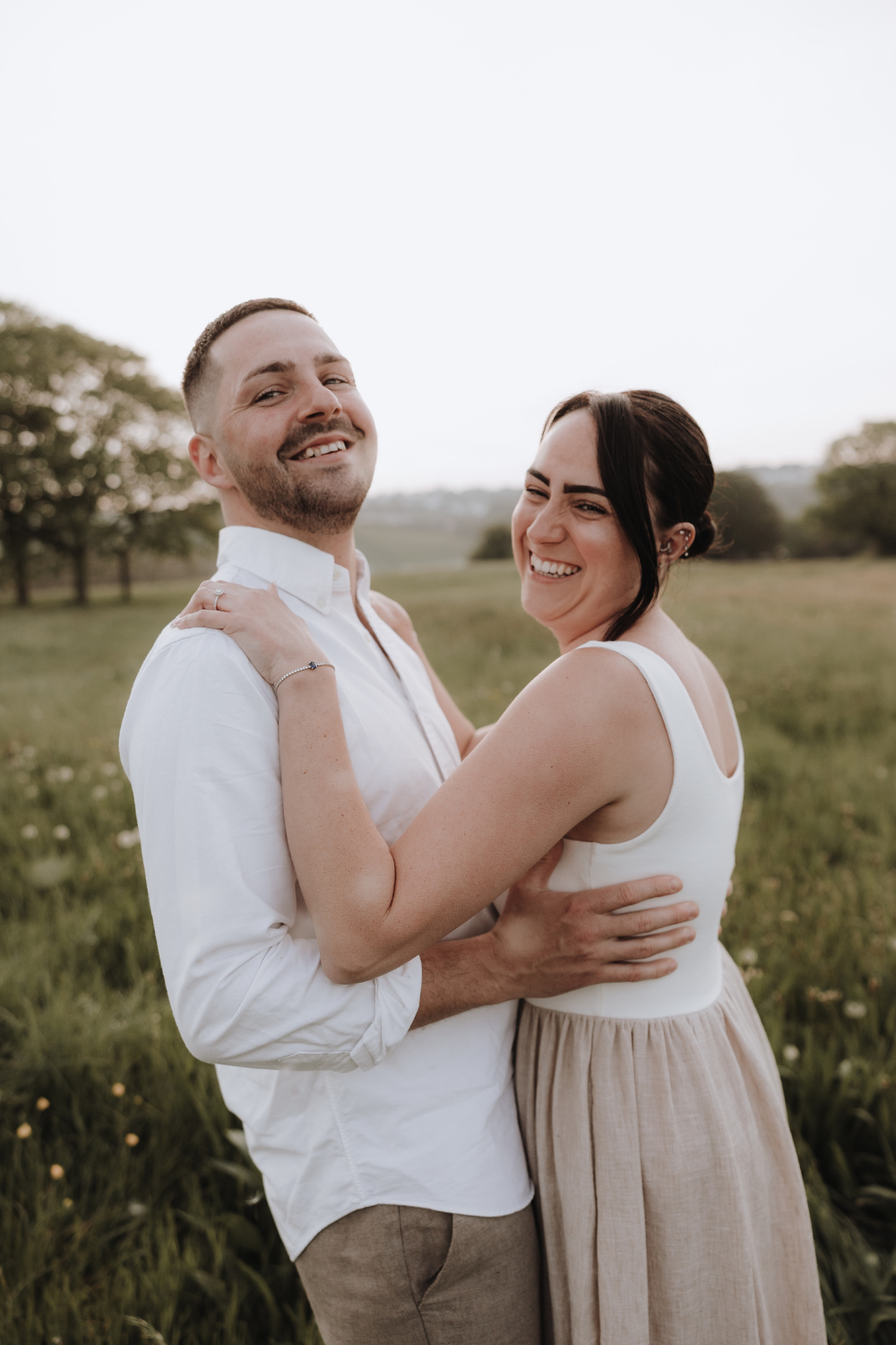 A smiling couple embraces in a lush green field, the man in a white shirt and woman in a white top and tan skirt.