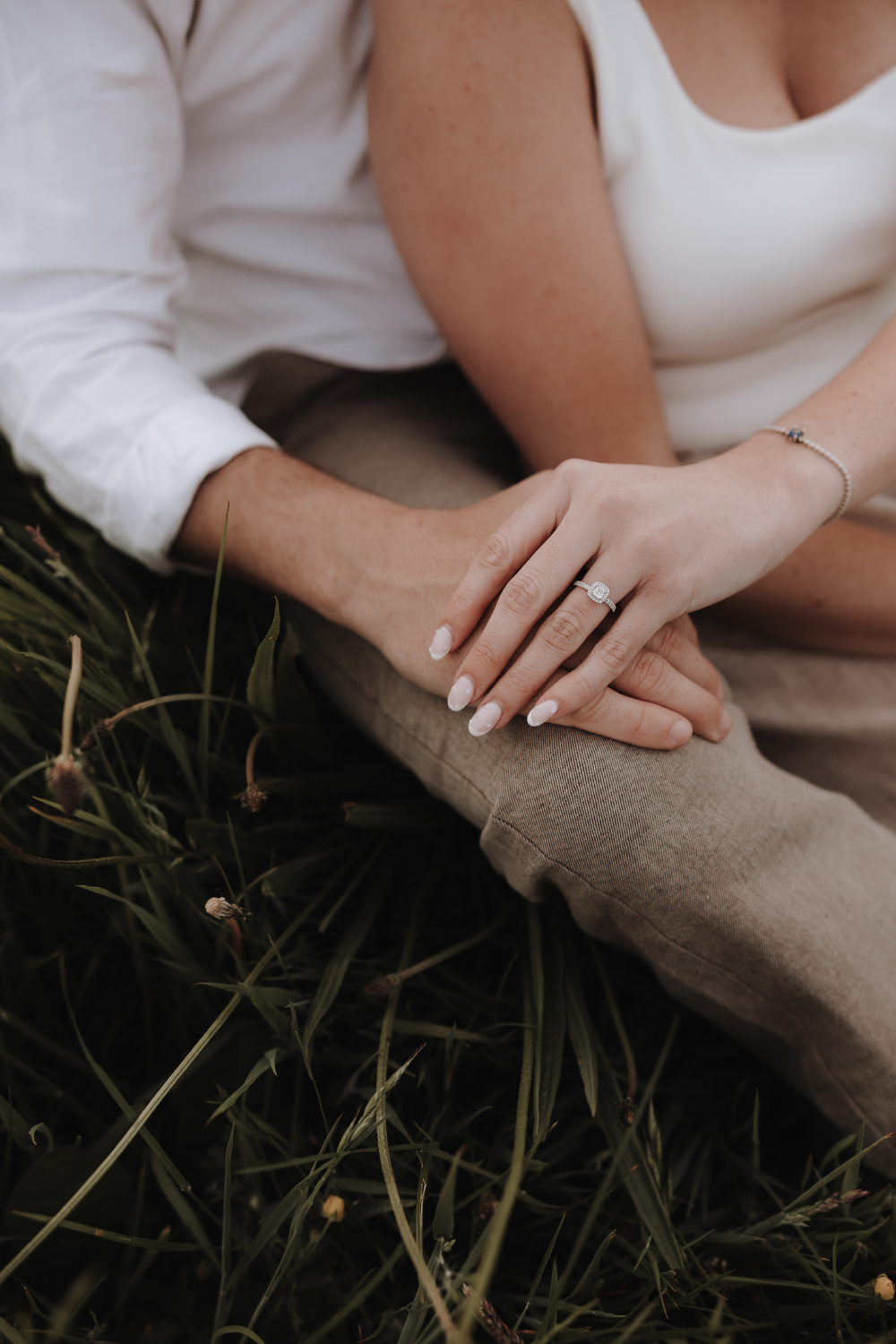 Close-up of couple sitting in grass, woman's hand with engagement ring resting on man's knee.