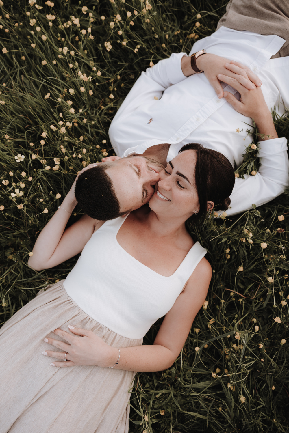 Couple lying in a wildflower meadow, sharing a tender kiss surrounded by green grass and small yellow flowers.