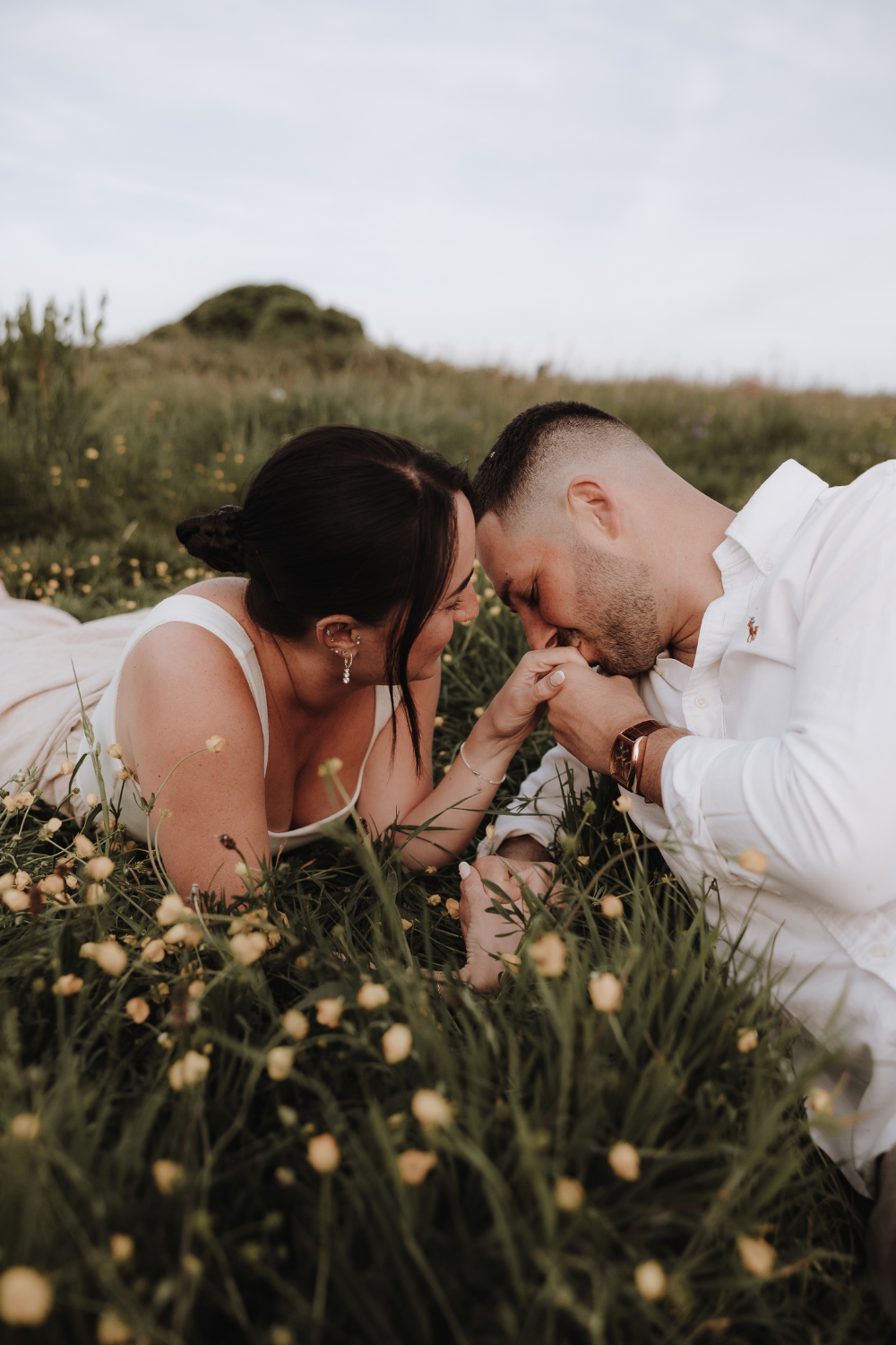 A romantic couple lies in a wildflower field, the man kissing the woman's hand as they share an intimate moment.