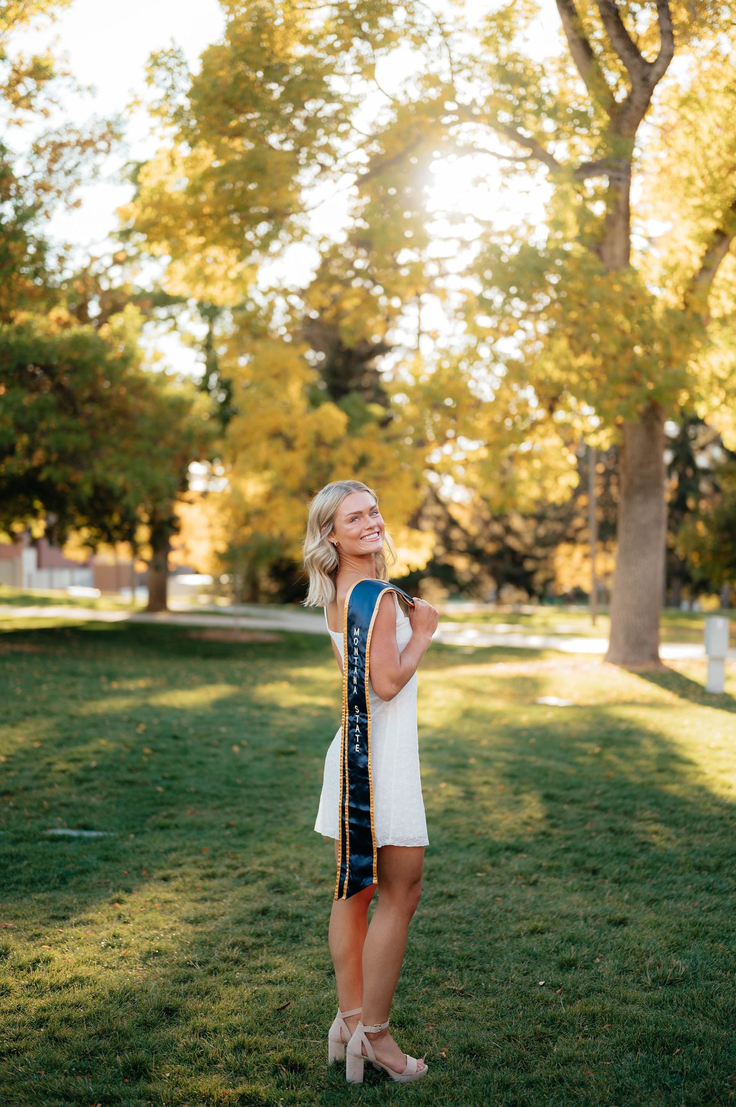 Blonde woman in white dress with graduation sash standing on green lawn with golden autumn trees.