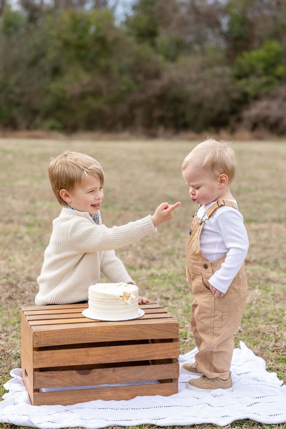 Two young children in beige and white outfits interact playfully while sitting on a wooden bench outdoors.