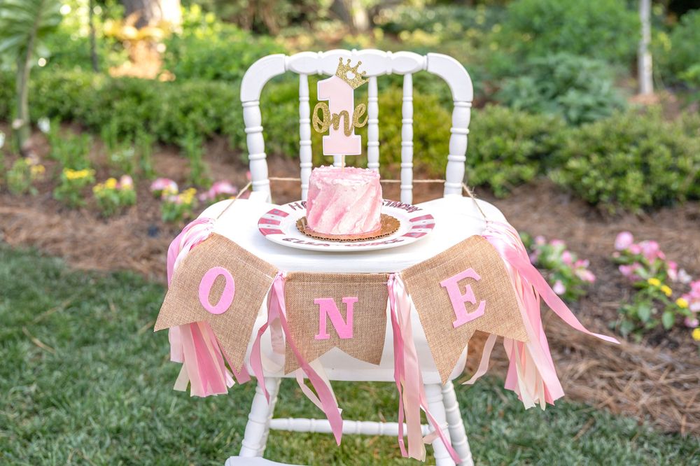 Pink and gold first birthday party decorations setup in garden with cake, highchair, and banner for outdoor celebration.