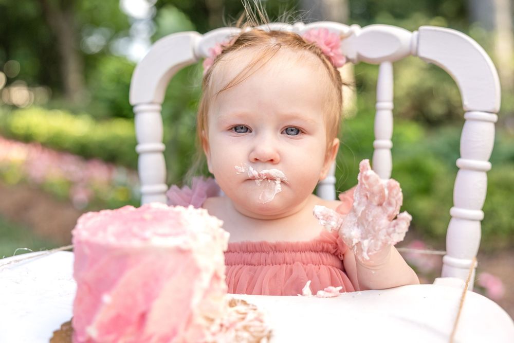 A baby in a pink dress sits on a white wooden high chair for her Raleigh NC cake smash at WRAL Azalea Gardens.