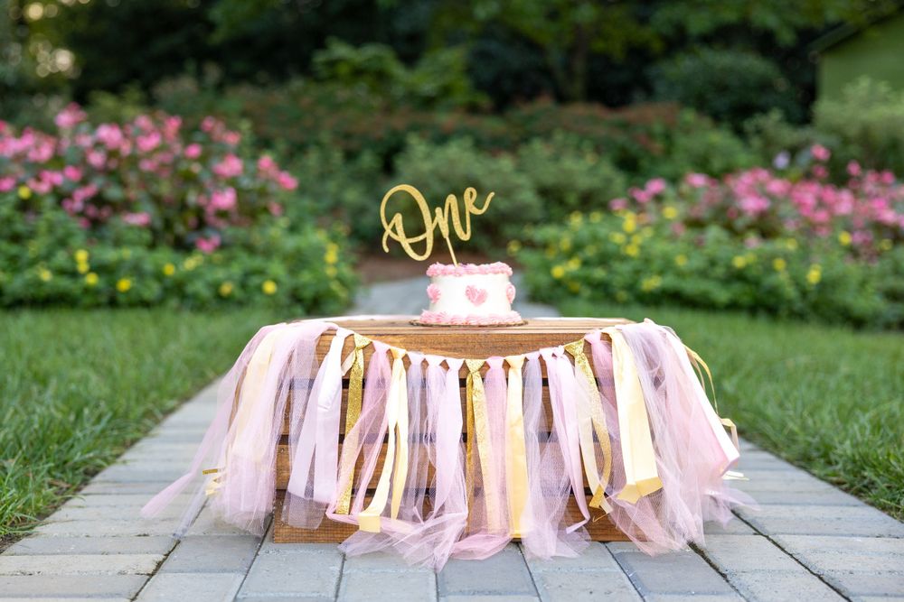 Pink and gold decorative cake smash setup with fabric tassels and 'One' topper on wooden deck surrounded by garden flowers.