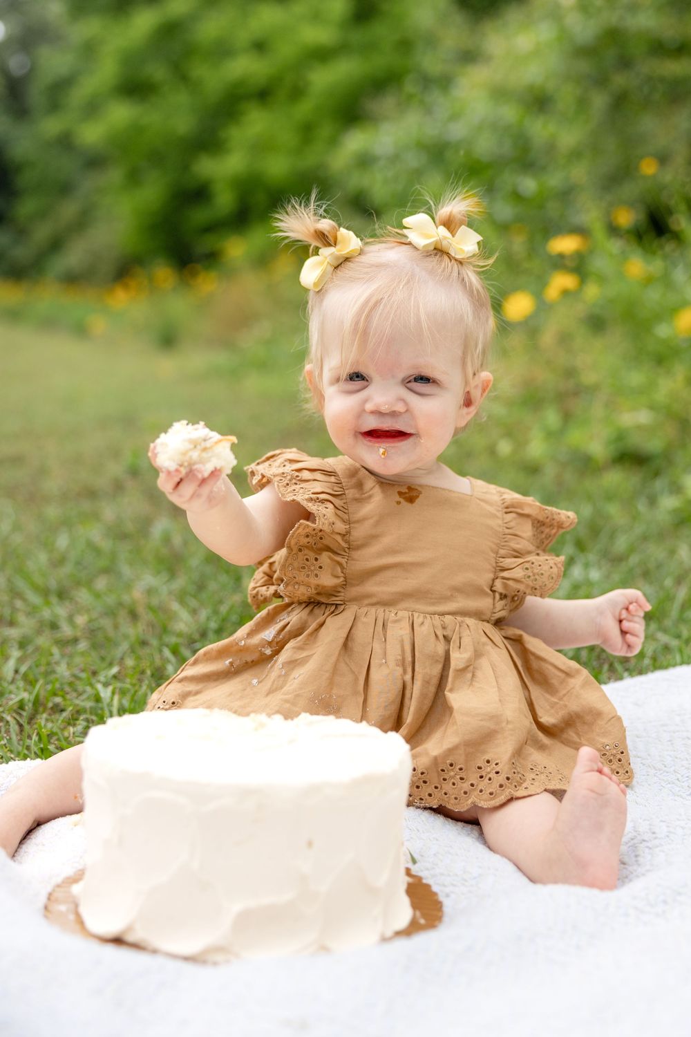 Small child wearing brown ruffled dress sits on white blanket with birthday cake during her Raleigh outdoor photo session.