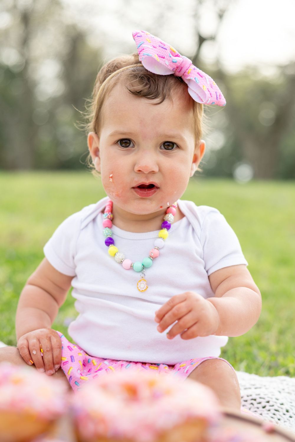 Baby in white shirt wearing pink bow and colorful necklace sits on blanket outdoors in grass.
