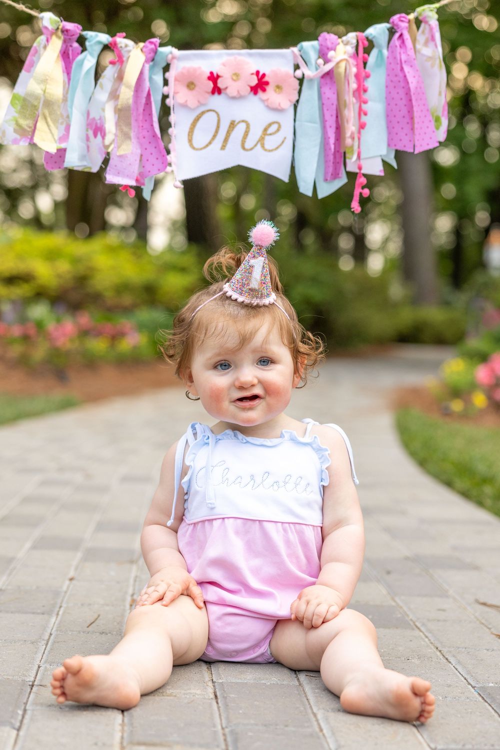 A baby wearing a pink outfit sits on stone pavers under a decorative banner with soft bokeh background for her First birthday photos in NC