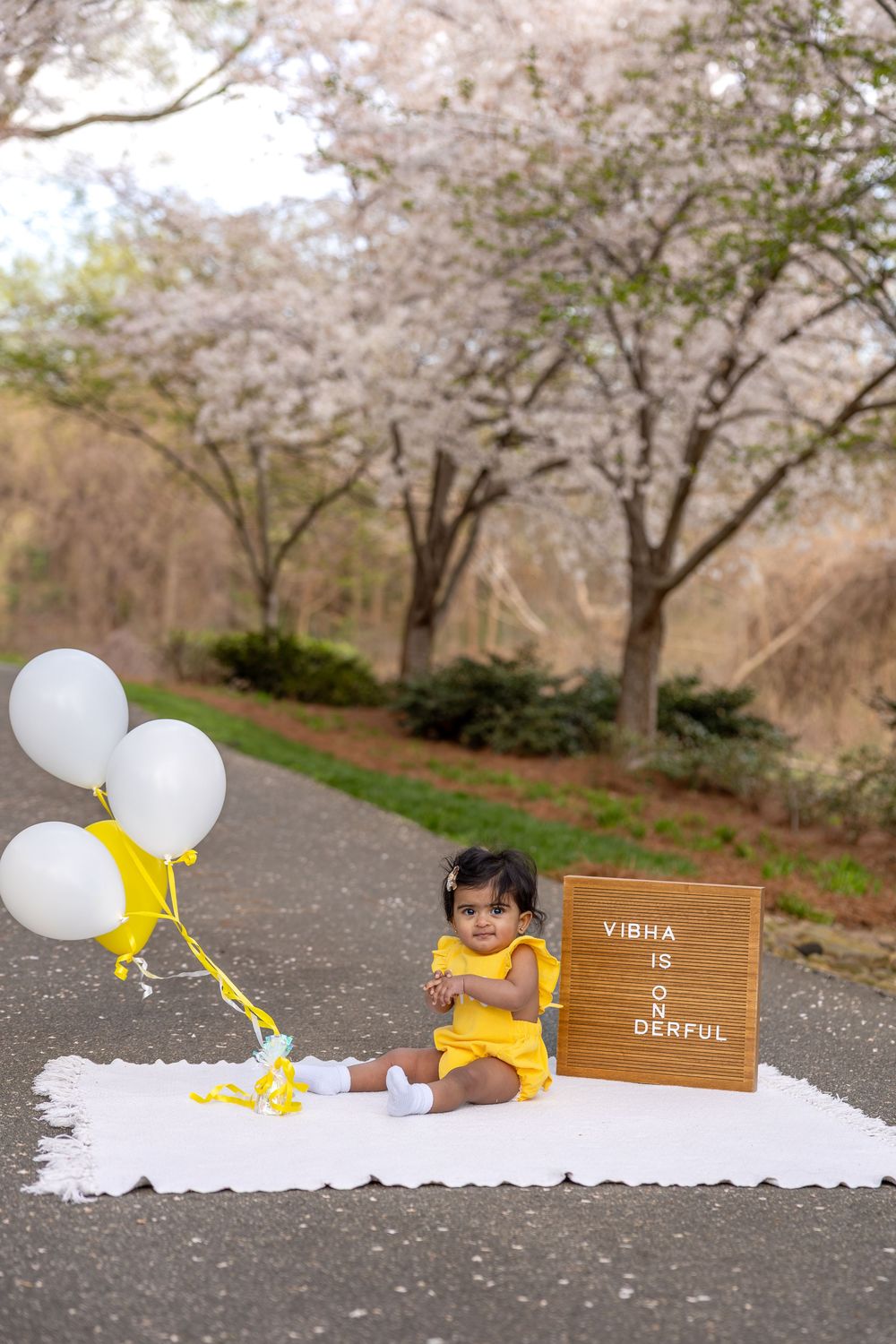 Baby girl in yellow dress sits on white blanket outdoors with white balloons - first birthday cake smash in Raleigh