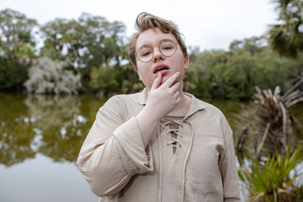 A person in a beige sweater poses thoughtfully by a serene lake surrounded by trees.