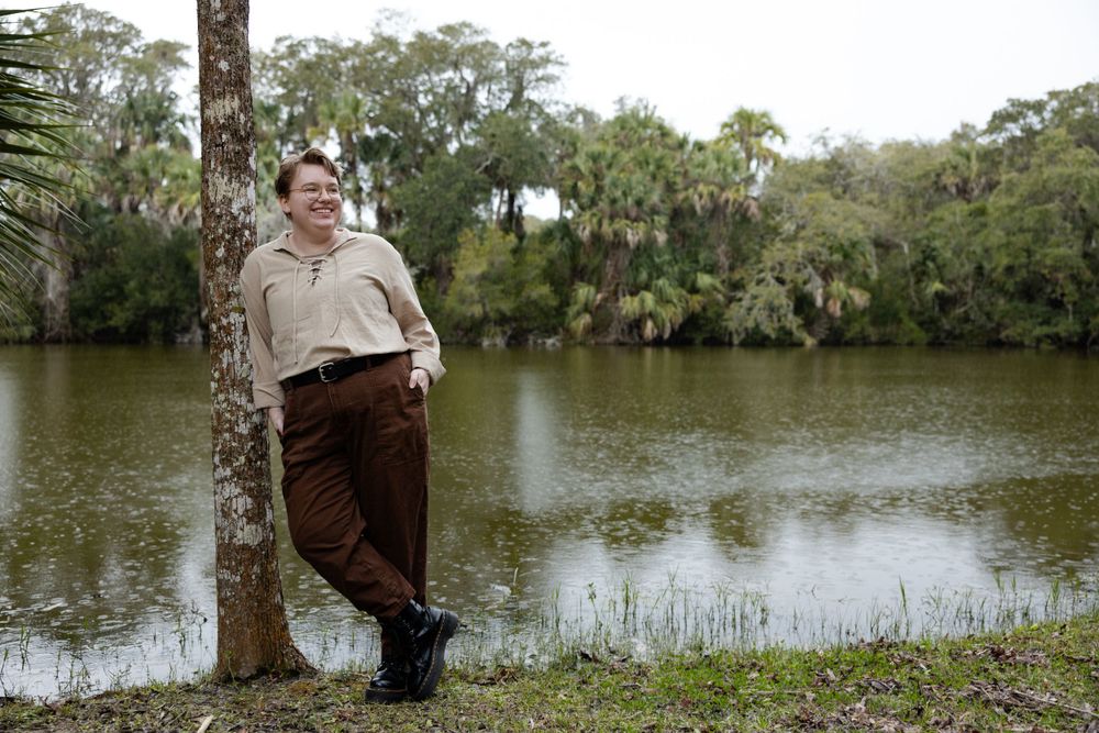 A figure in casual wear leans against a tree beside a tranquil lake in a wooded setting.