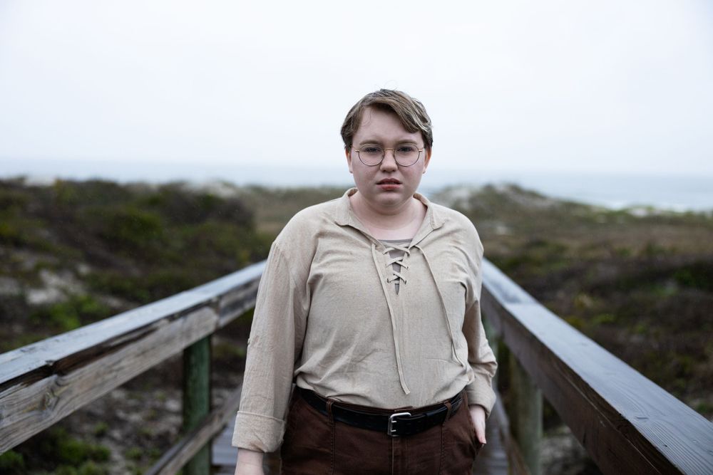 A person in a beige top stands on a wooden boardwalk with natural scenery in the background.