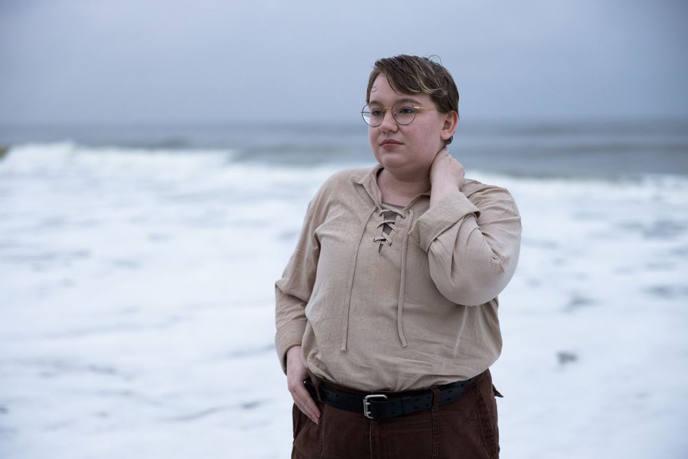 A person in brown pants and beige sweater stands against a dramatic ocean backdrop.