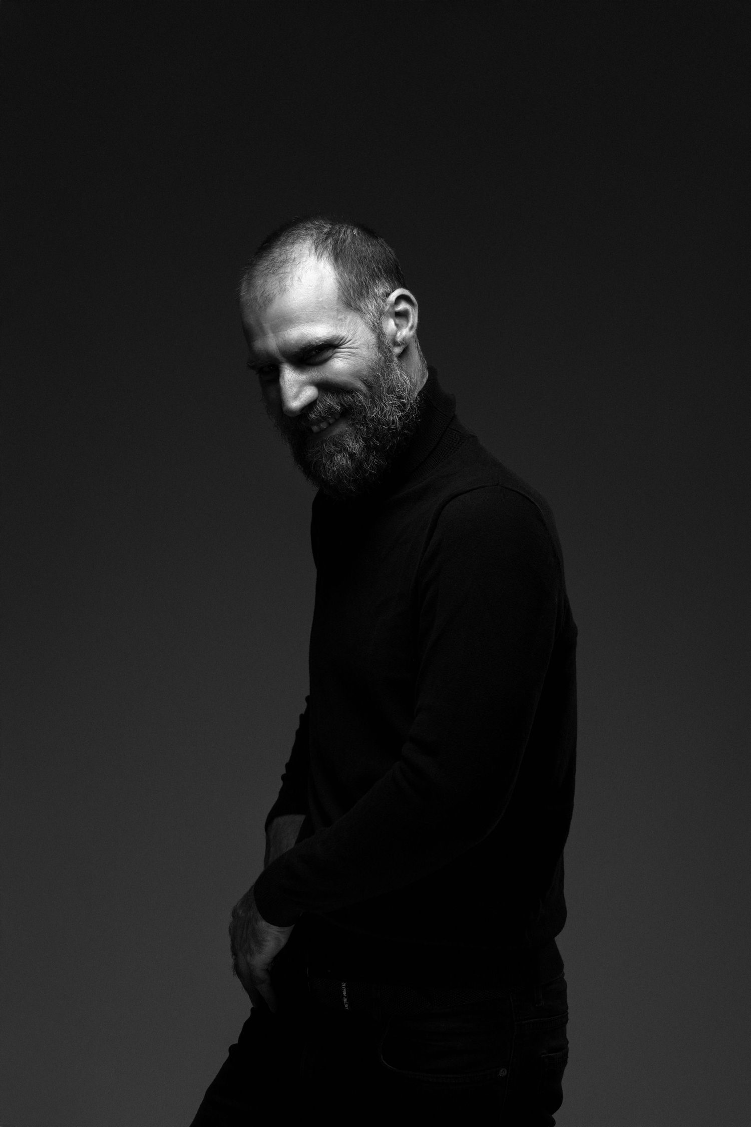 Dramatic black and white portrait of Jose Manuel Poga wearing a black turtleneck against a dark background.