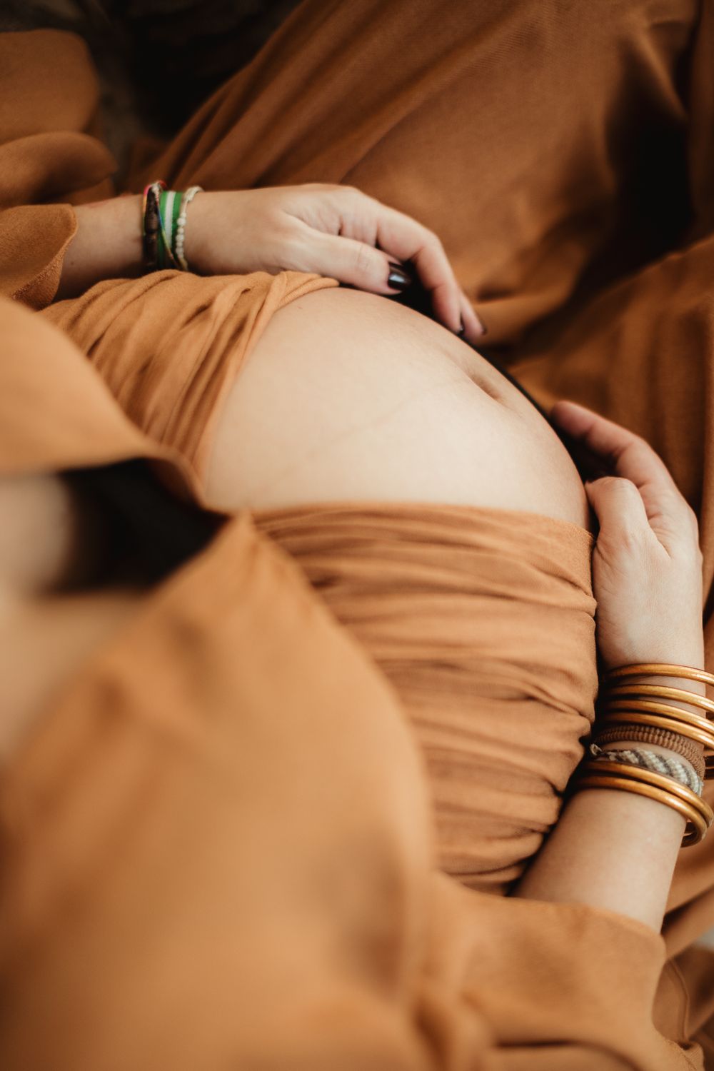 Close-up of hands with bracelets resting on a pregnant belly wrapped in warm brown fabric.