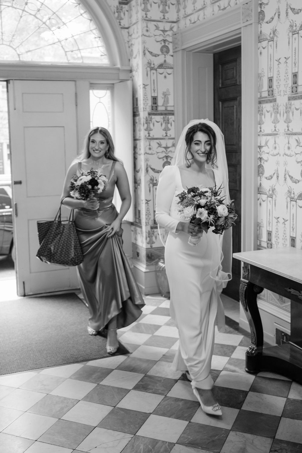 Black and white photo of a bride and bridesmaid walking in an elegant hallway with checkered floors and ornate walls.