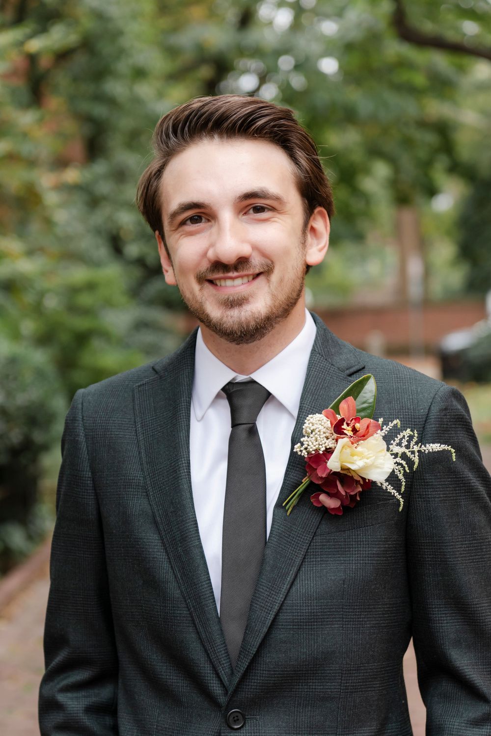 Groom in a green suit smiles outdoors wearing burgundy and cream boutonniere with seasonal greenery.