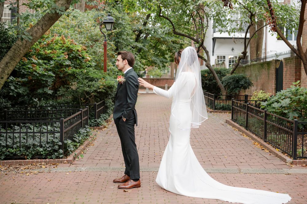 Bride and groom share a first look moment on a brick pathway surrounded by lush greenery and iron railings.