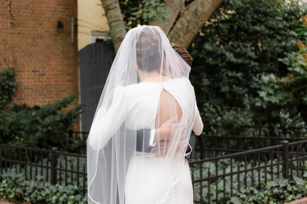 A bride in a white wedding dress and flowing veil gets a hug from her groom during their first look.