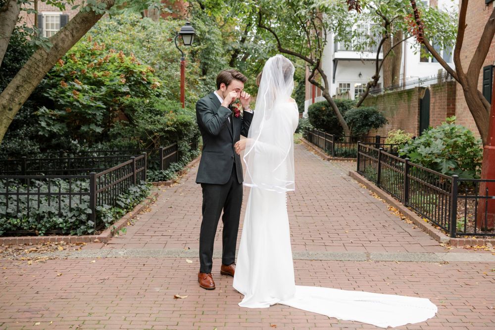 A groom wipes his tears during a romantic moment on a brick path surrounded by lush greenery in an urban garden setting.