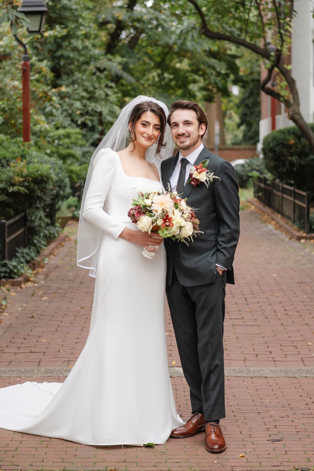 Wedding couple stands on brick walkway with trees, bride holding bouquet of pink and white flowers in this timeless portrait.