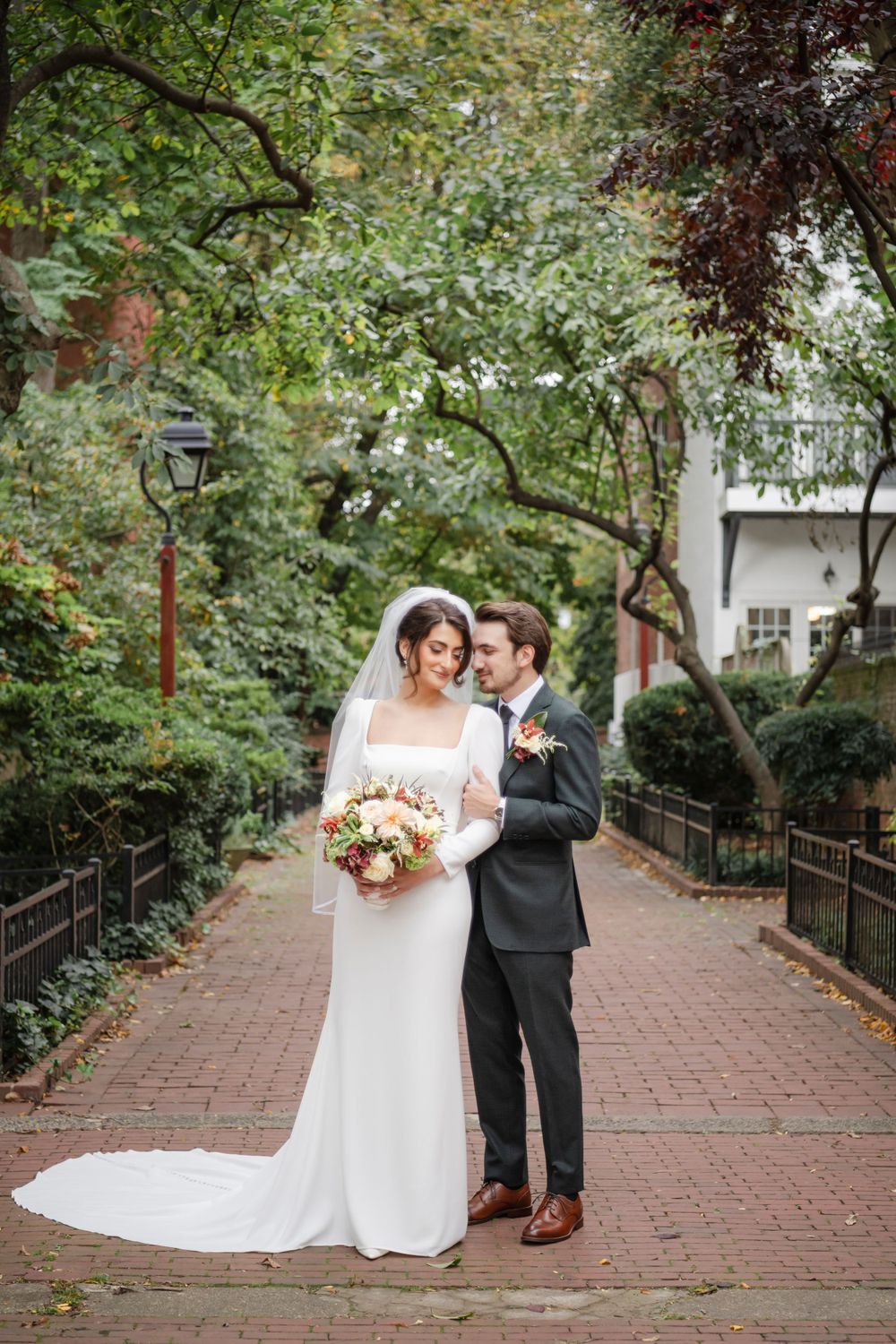 A wedding couple stands on a brick path surrounded by lush greenery and trees in a romantic garden setting.