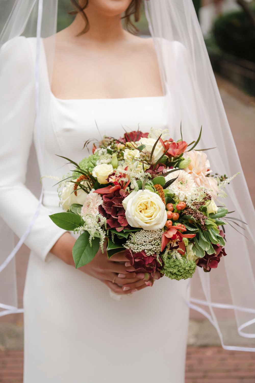 Elegant wedding bouquet with cream roses, burgundy flowers and greenery held against white long-sleeved bridal gown.