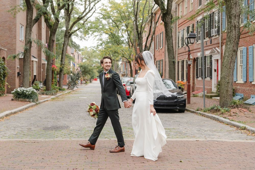 A couple in wedding attire walks down a charming cobblestone street lined with historic brick buildings and trees in Philadelphia
