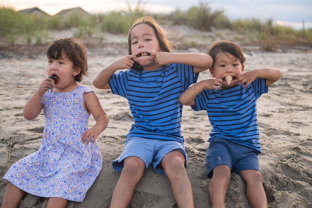 Three playful children making silly faces while sitting together on a sandy beach.