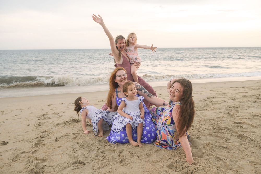 Joyful group in matching floral dresses playfully posing and laughing together on a sunny beach at sunset.