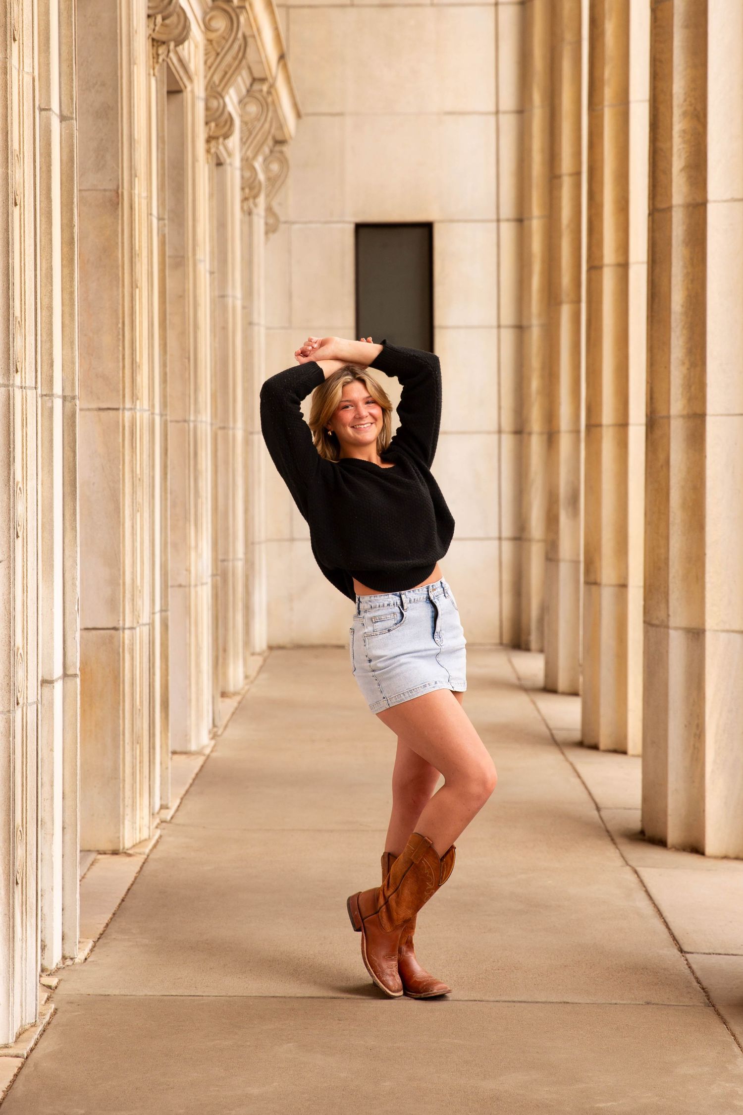 Smiling blonde senior in black sweater, denim shorts, and brown cowboy boots posing between stone columns.