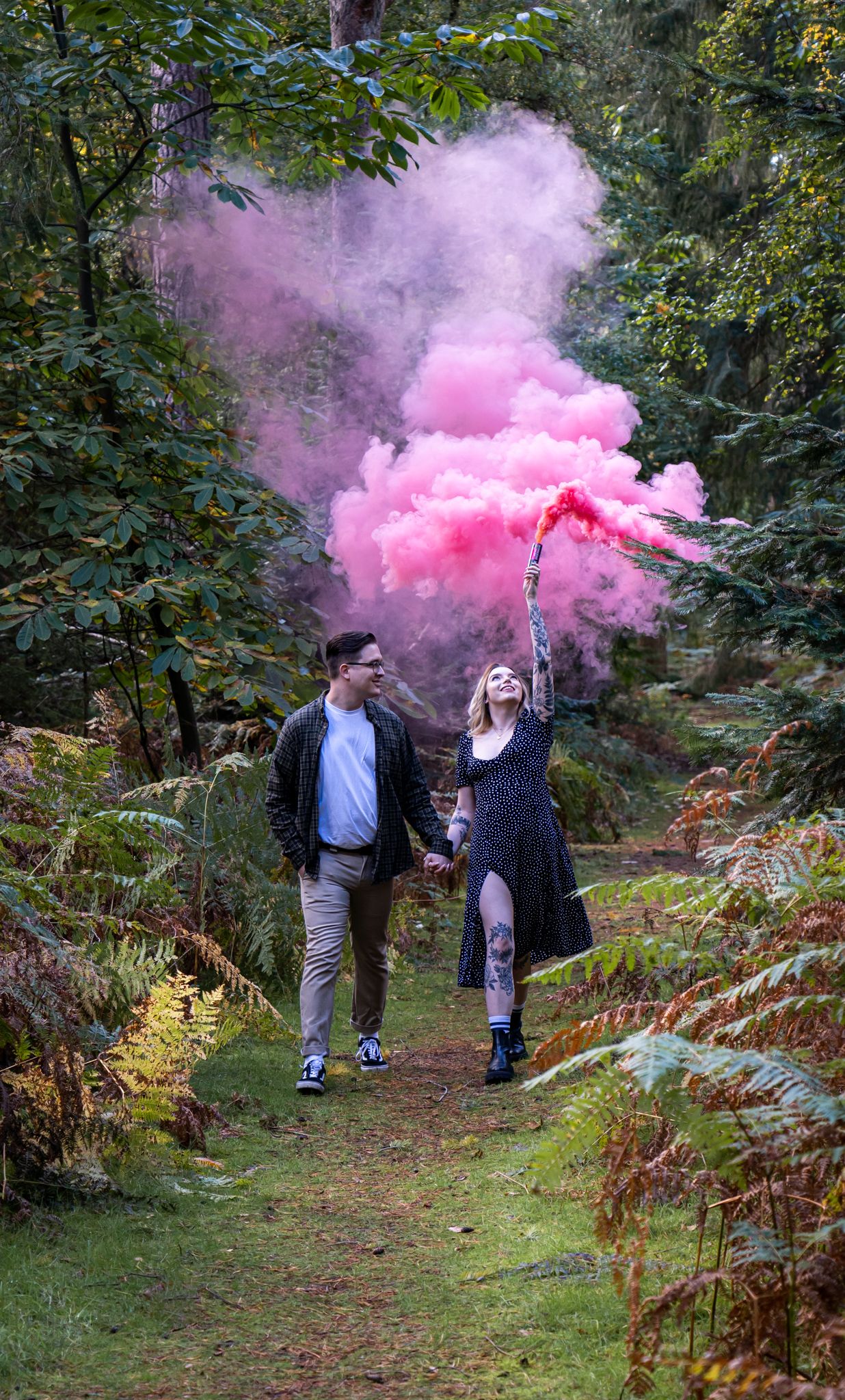 Couple holds pink smoke flares while walking through mystical forest path.