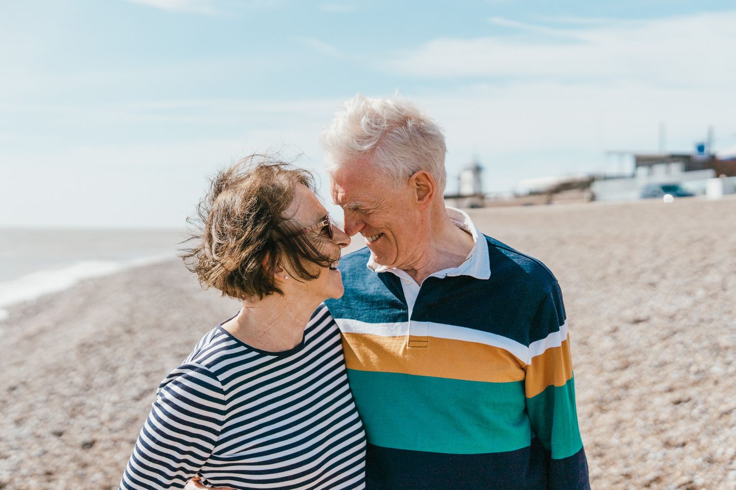 Happy couple sharing an intimate moment on a sunny beach, wearing casual striped and colorful clothing.