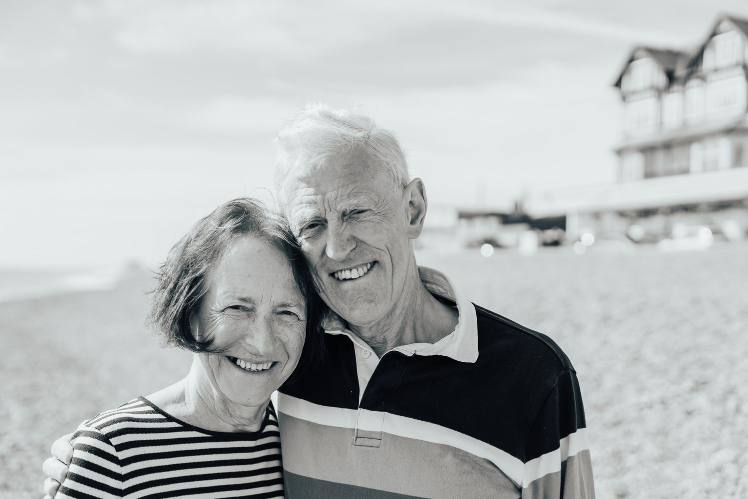 Black and white portrait of a mature couple laughing together on a beach with houses in background.